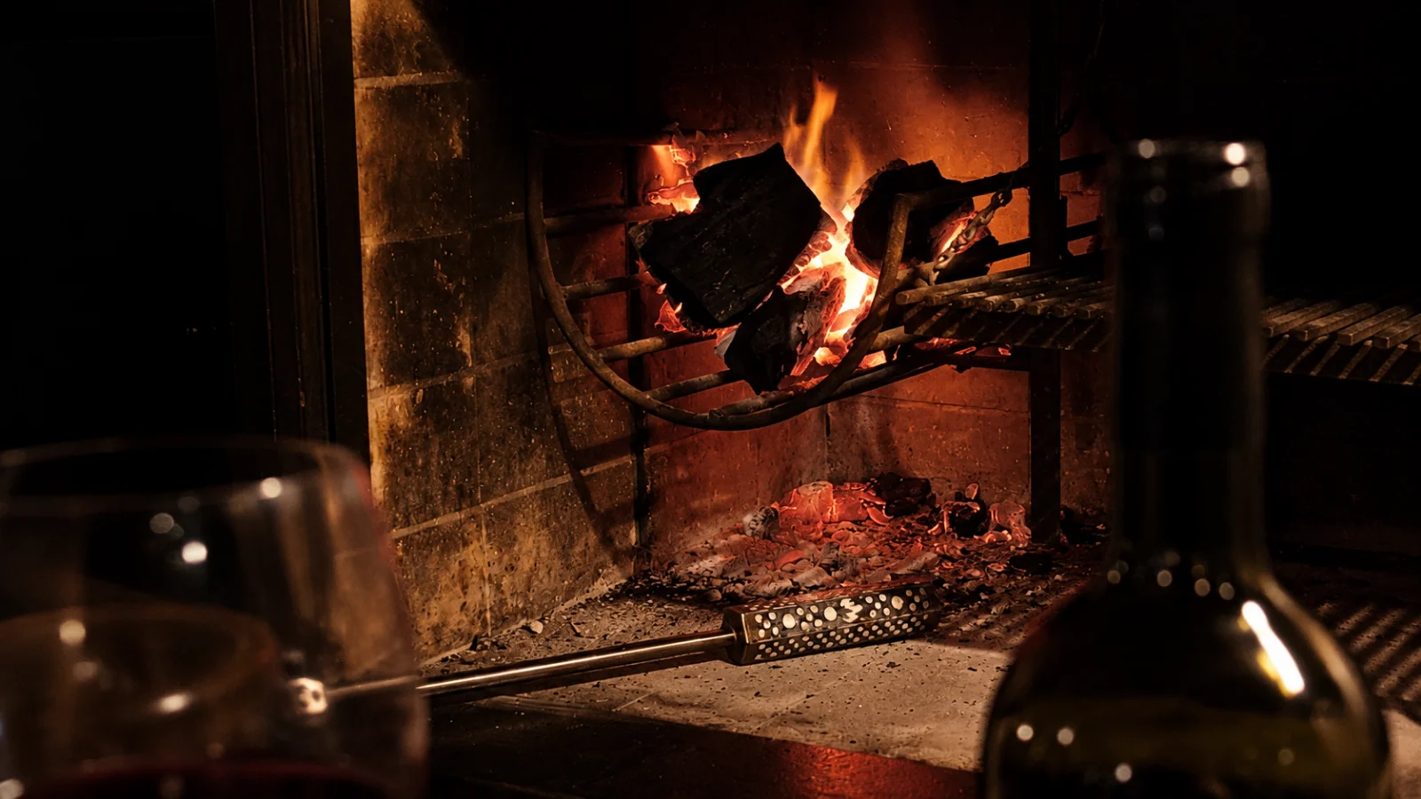 Fuego encendido en un hogar a leña, con el Iniciador de Fuego MIL ASADOS apoyado sobre las cenizas y una copa de vino en primer plano