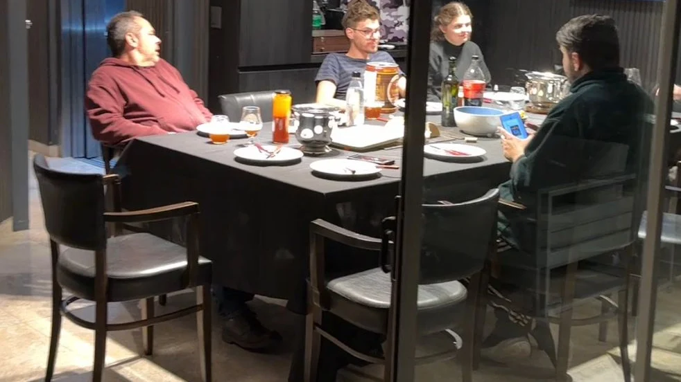 Una mesa larga vista desde afuera a través de un ventanal, con varias personas sentadas alrededor en una cena de noche